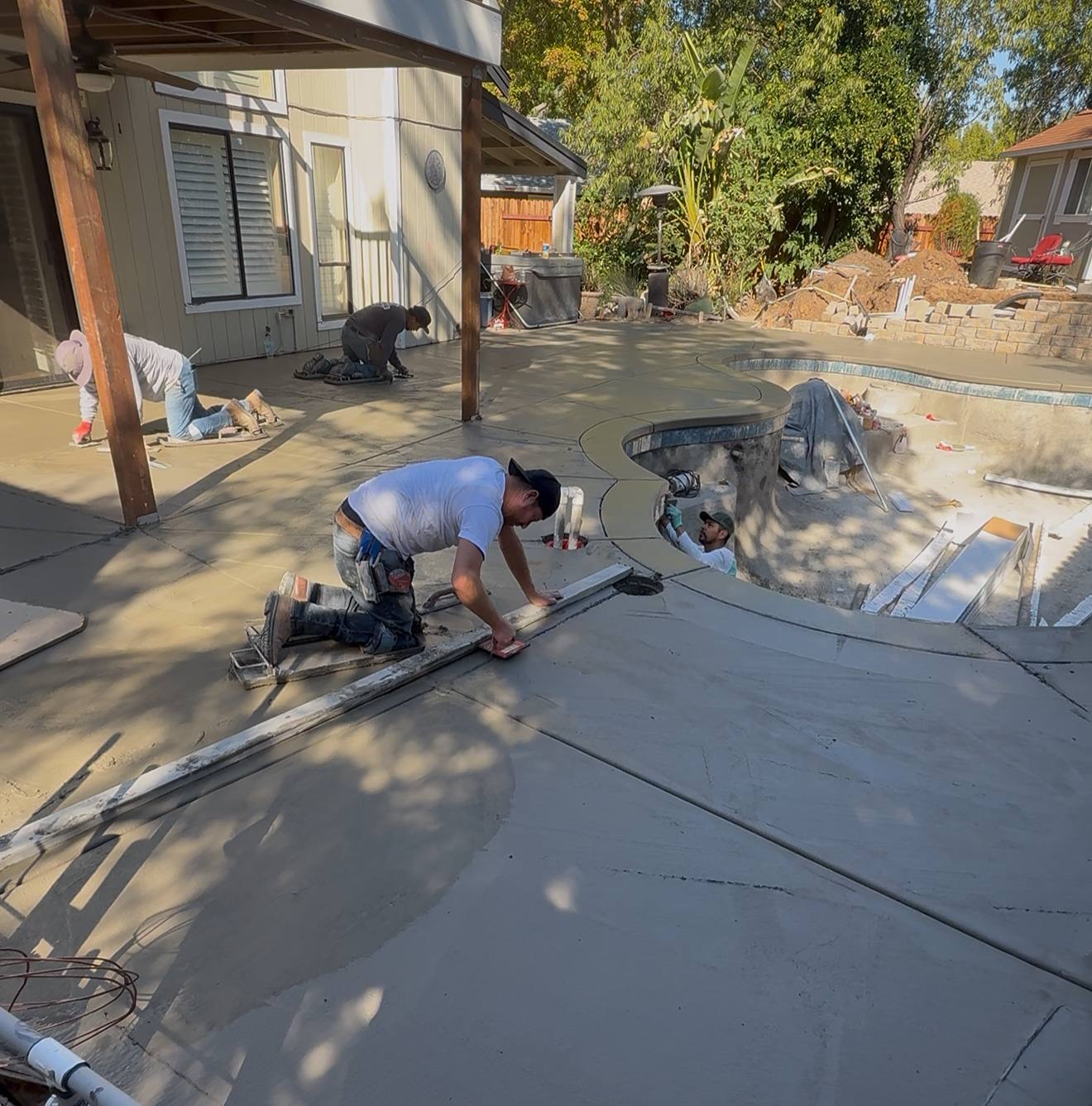 Crew finishing a new concrete deck around a pool under a wooden patio cover